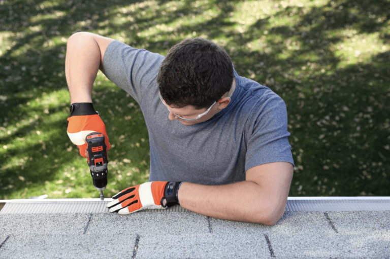 A person wearing safety glasses and orange gloves uses a power drill to install a Gutterglove stainless steel micro-mesh guard on the edge of a house roof. Green grass and shadows are visible in the background.